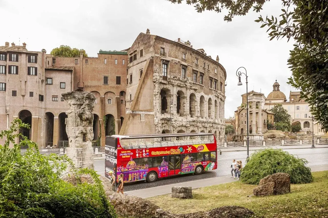 Un bus touristique rouge vif, orn&eacute; d'images color&eacute;es de monuments, circule devant des ruines antiques. Un excellent moyen de faire un rome bus tour !