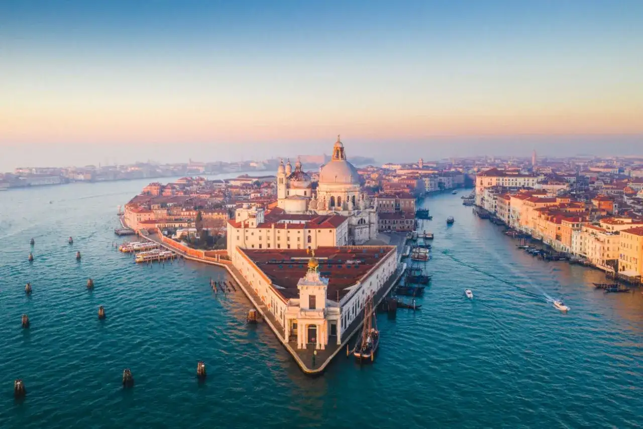 Vue aérienne de la Dogana de Venise, un bâtiment triangulaire emblématique au bord du Grand Canal, baigné par la lumière dorée du soleil couchant.