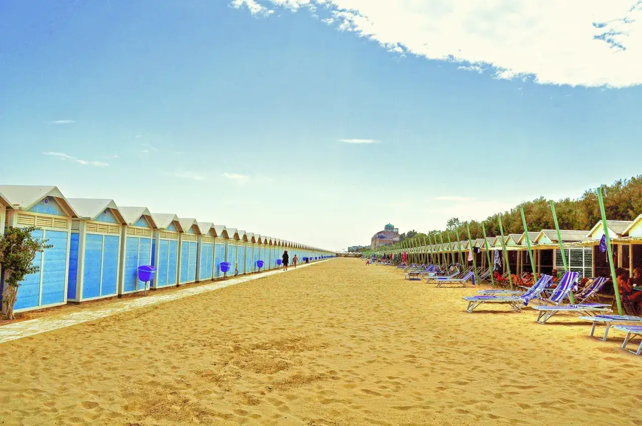 Plage de sable doré au Lido di Venezia, avec des cabines bleues alignées d'un côté et des chaises longues sous des auvents de l'autre.