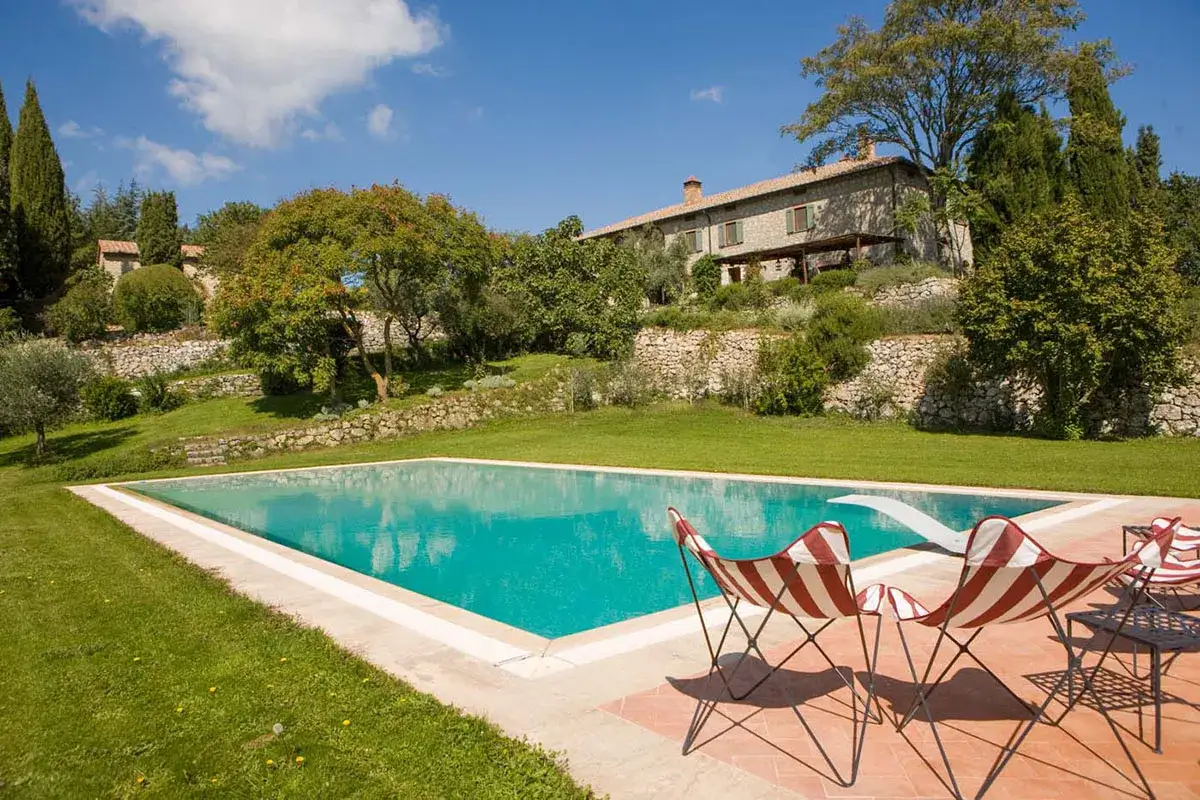 Une magnifique piscine dans un cadre toscan, avec une maison en pierre en arrière-plan et des chaises rayées invitant à la détente.
