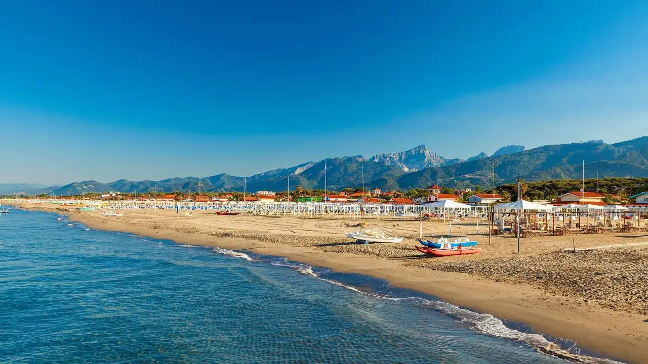 Plage de sable fin à Tirrenia, Italie, avec des rangées de parasols blancs, des montagnes en arrière-plan et la mer bleue.