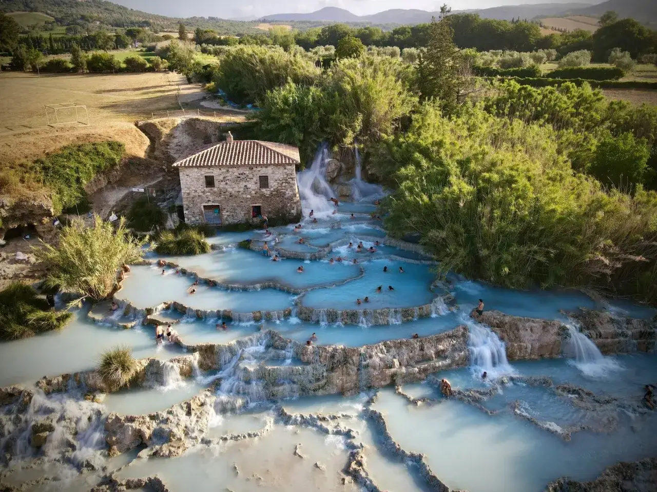 Cascades d'eau thermale bleue &agrave; Rapolano Terme, avec une vieille b&acirc;tisse en pierre et des baigneurs profitant de la d&eacute;tente.