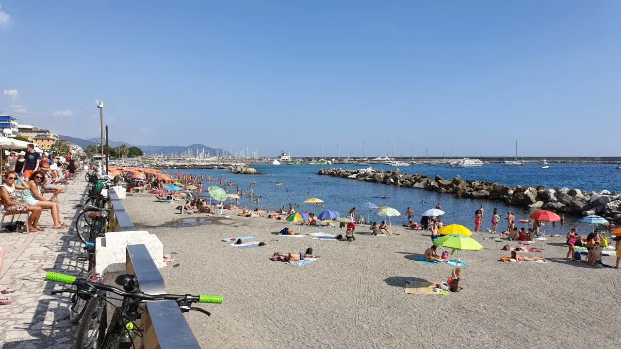Plage animée à Chiavari, baigneurs profitant du soleil sous des parasols colorés, avec des bateaux dans le port.
