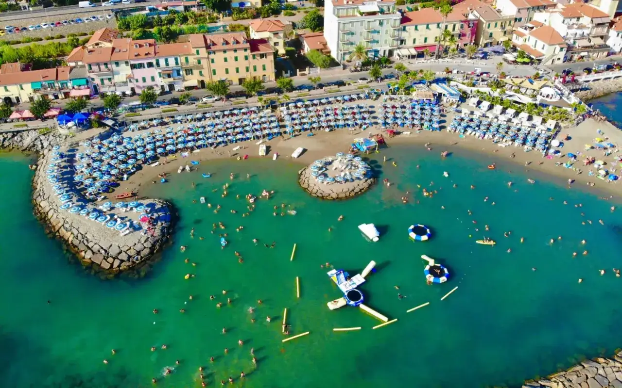 Plage animée à Arma di Taggia, Italie, avec des parasols bleus et blancs, des baigneurs dans la mer turquoise et des structures gonflables.
