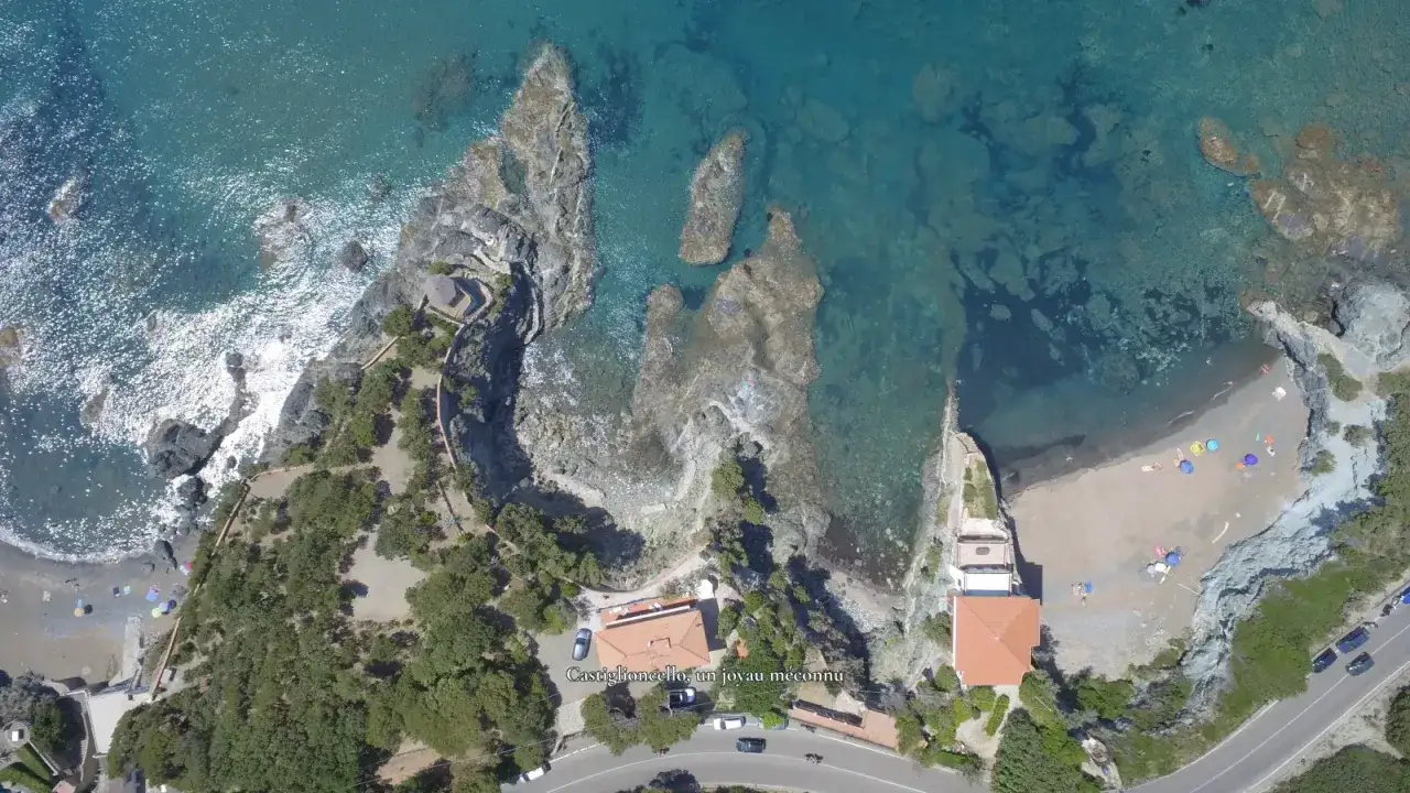 Vue aérienne de Castiglioncello, Italie. La côte rocheuse rencontre une plage de sable avec des parasols colorés. L'eau turquoise révèle les fonds marins.