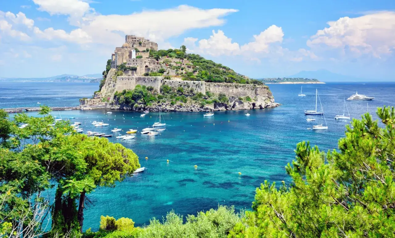 Vue d'un château médiéval sur une île verdoyante, baignée par les eaux turquoise de la baie de Naples, avec des voiliers et des yachts.