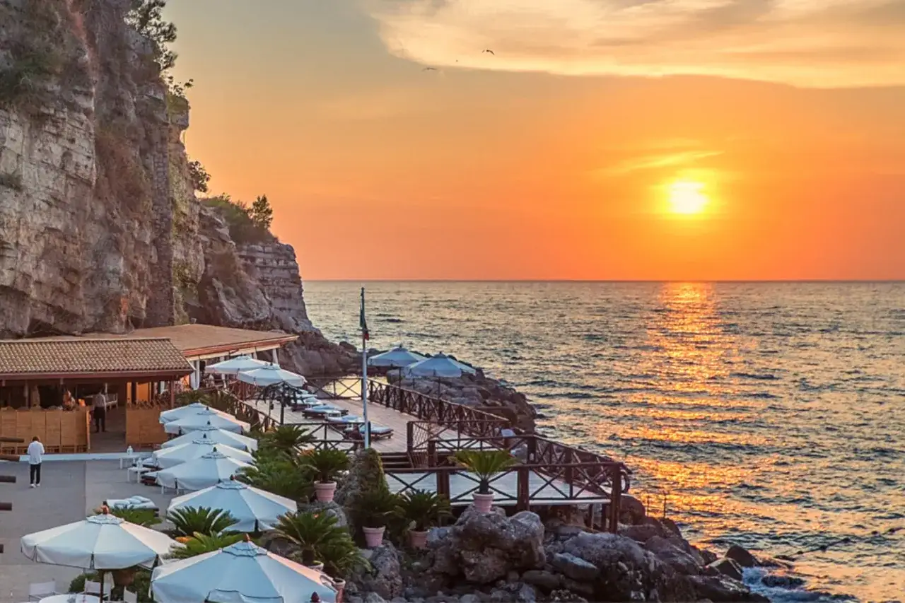 Coucher de soleil sur la mer à Vico Equense, avec une falaise rocheuse, un restaurant et des parasols blancs.