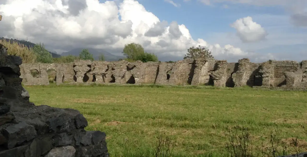 Vestiges d'une ancienne structure en pierre en Italie, avec des arches et des ouvertures sur un champ verdoyant sous un ciel nuageux.