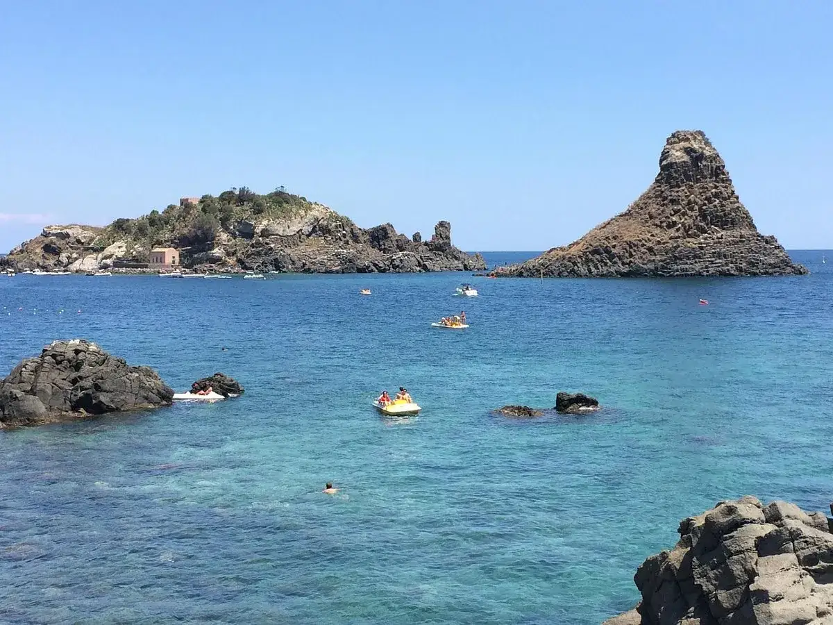 Vue sur la mer bleue cristalline à Aci Trezza, Sicile, avec des îlots rocheux et des bateaux.