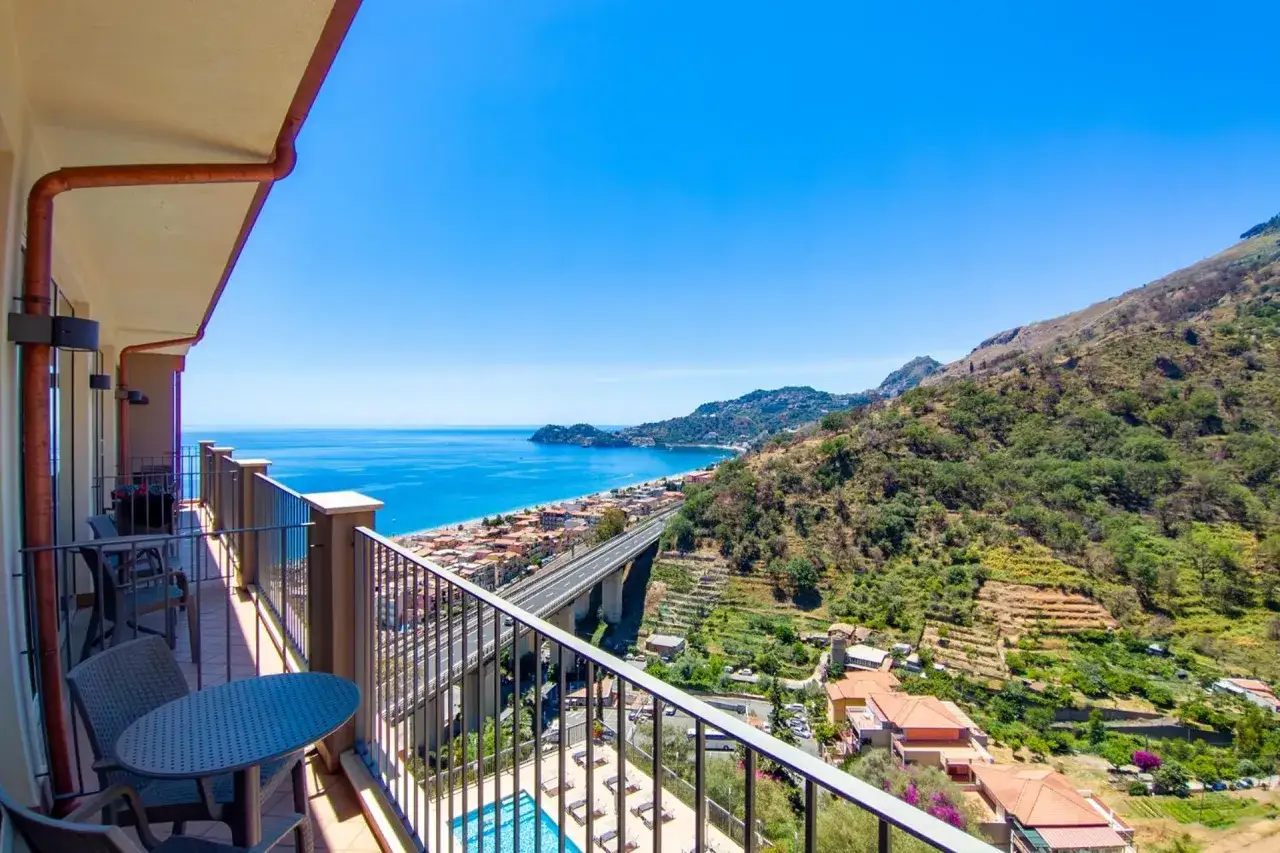 Vue depuis le balcon de l'hôtel Antares Sicile : mer bleue, ville côtière, pont autoroutier et colline verdoyante.