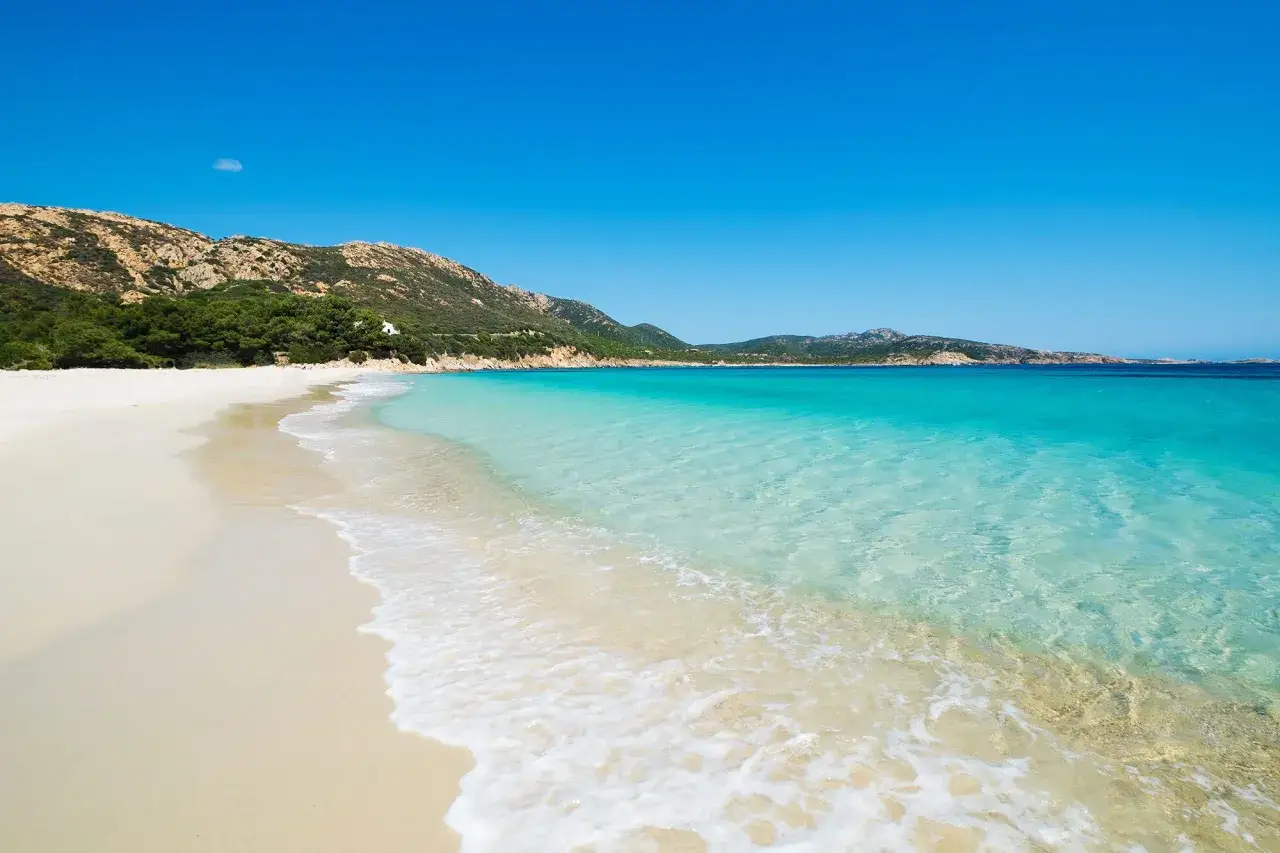 Plage de sable blanc bordée par une eau turquoise cristalline, avec des collines verdoyantes en arrière-plan. Une destination de rêve en Italie.