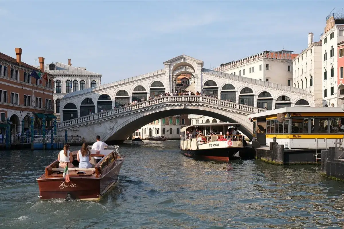 Bateaux naviguant sur le Grand Canal sous le célèbre Ponte Rialto, avec des passagers profitant du soleil vénitien.