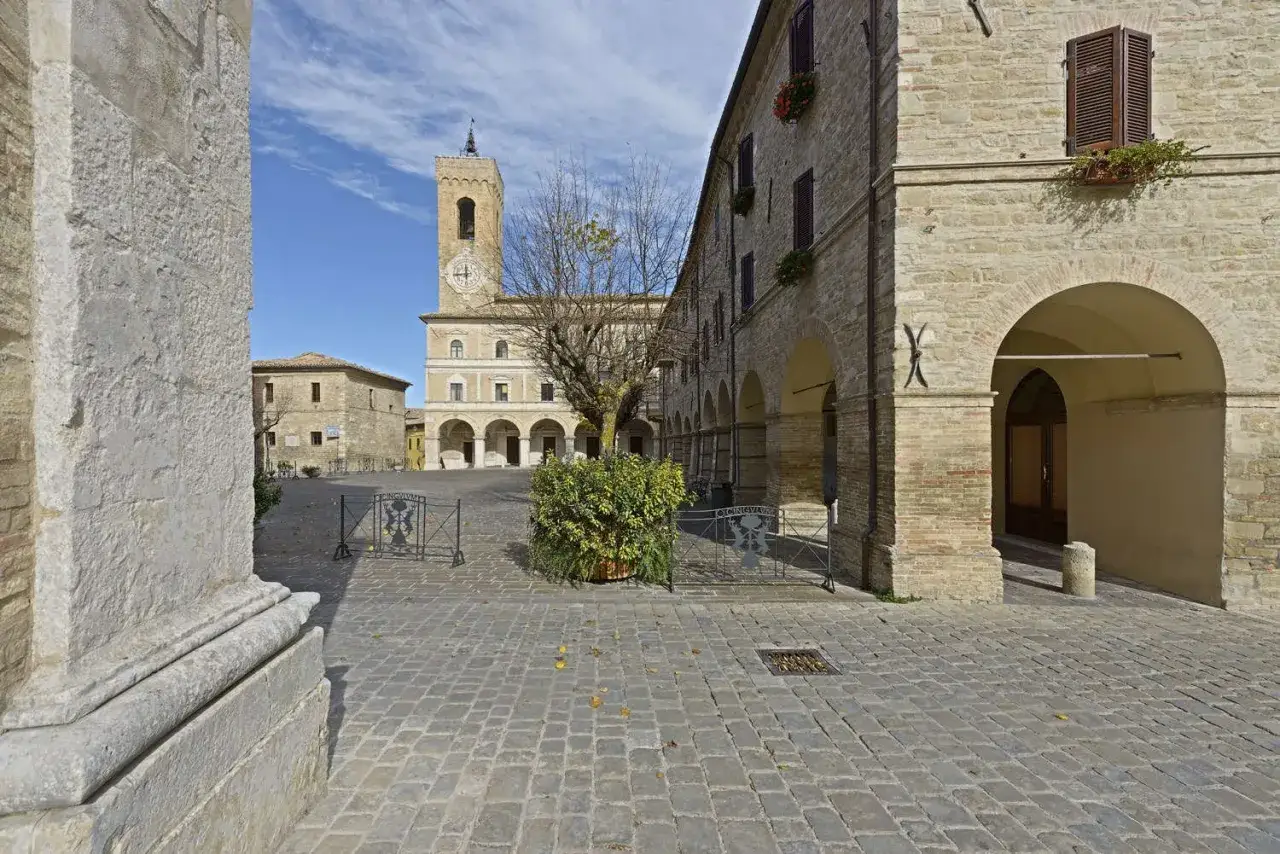 Place pav&eacute;e de Cingoli avec une tour d'horloge imposante, des arcades et des b&acirc;timents en pierre sous un ciel bleu.