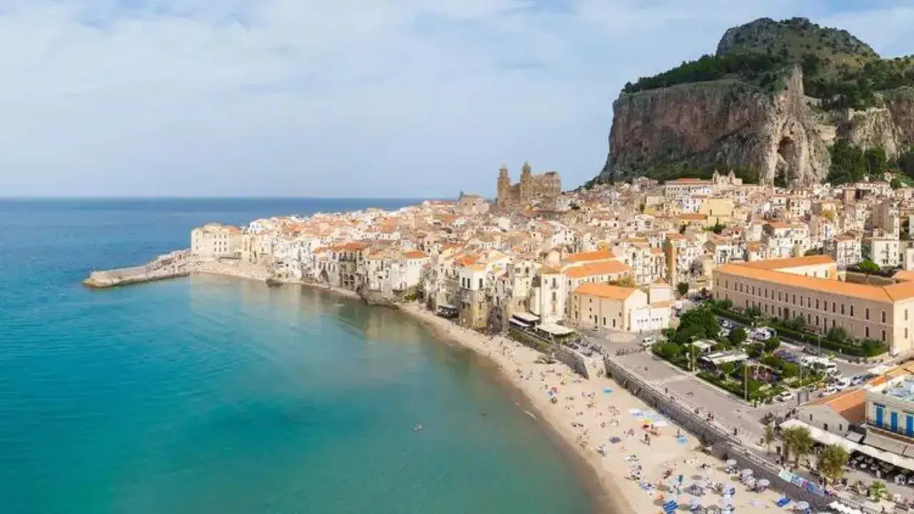 Vue aérienne d'une ville pittoresque en Italie, bord de mer, avec une plage animée et une mer turquoise. Un rocher imposant domine le paysage.