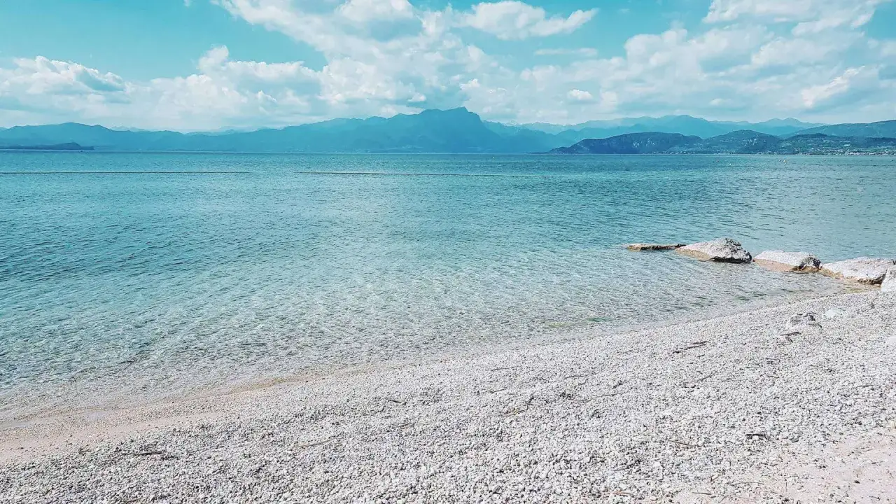 Plage de galets blancs au bord du lac de Garde, eaux bleues transparentes, montagnes bleues à l'horizon sous un ciel nuageux.