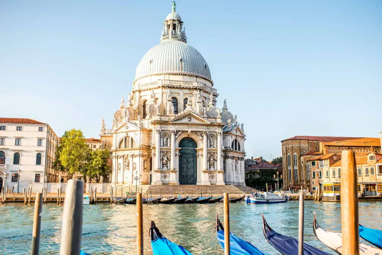 La majestueuse basilique Santa Maria della Salute à Venise, baignée de soleil, se reflète dans le Grand Canal. Des gondoles bleues et blanches attendent paisiblement.