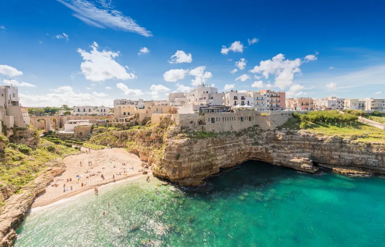 Vue panoramique de Polignano a Mare, avec ses maisons blanches perchées sur des falaises rocheuses surplombant une plage de galets et une mer turquoise.