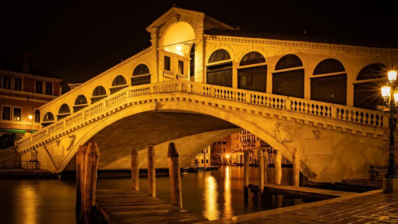 Le pont du Rialto illuminé la nuit, avec ses arches et ses boutiques, se reflétant dans les eaux sombres du Grand Canal.