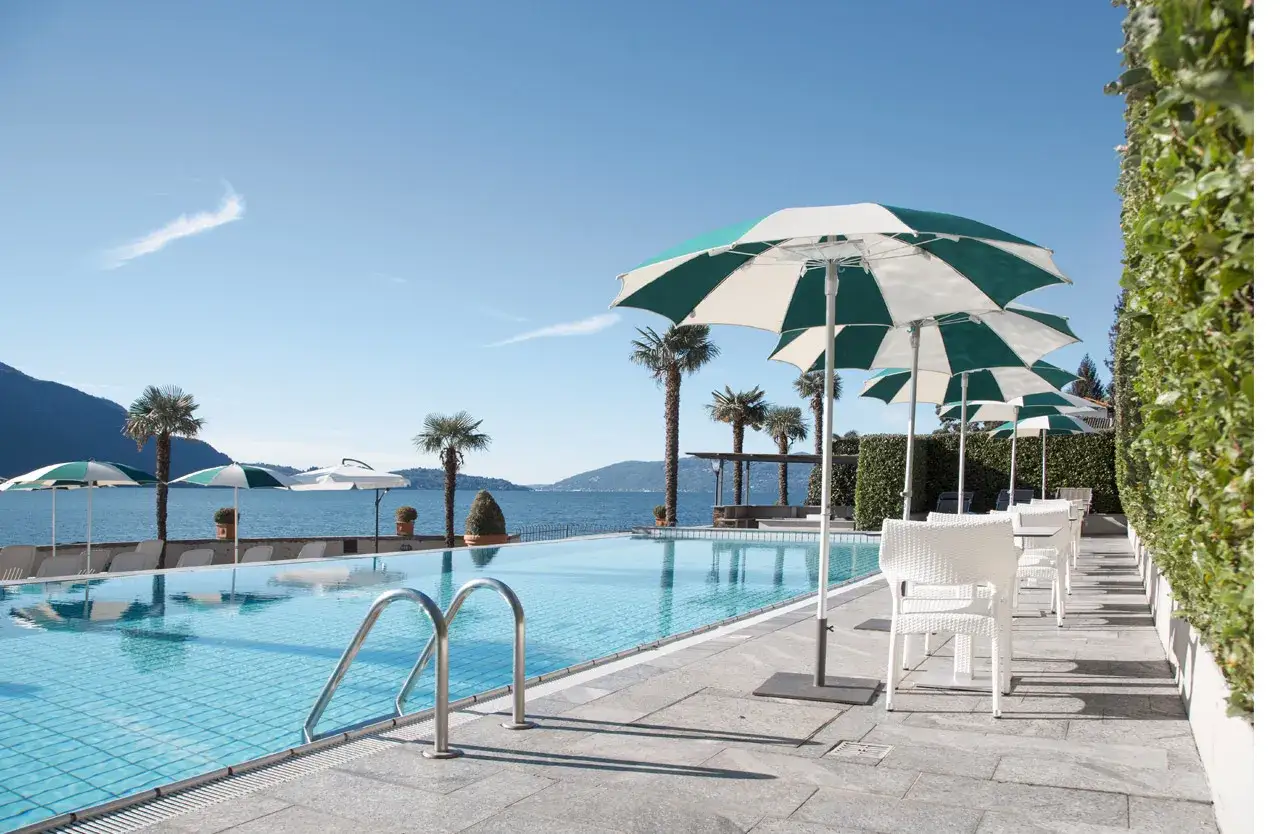 Piscine d'un h&ocirc;tel &agrave; Ghiffa sur le Lac Majeur, avec parasols, palmiers et vue sur le lac.