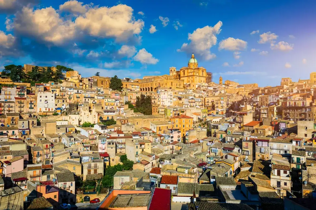 Vue panoramique de la ville de Piazza Armerina en Sicile, avec ses maisons colorées s'étageant sur une colline, dominée par une imposante église à dôme vert.