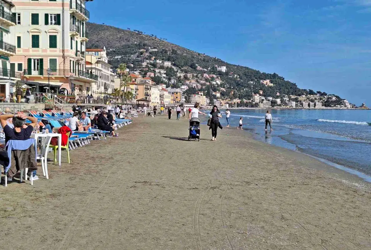 Plage animée à Alassio, Italie, avec des gens se relaxant sur des chaises longues et se promenant au bord de la mer, sous un ciel bleu.