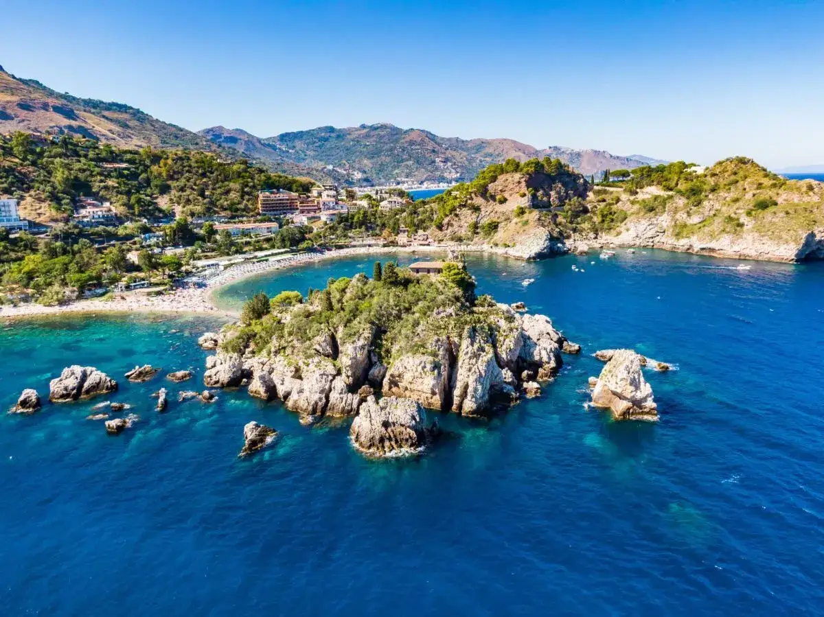 Île rocheuse verdoyante baignée par les eaux bleues de la mer, avec une plage animée et des collines en arrière-plan, près de Catane, Sicile.