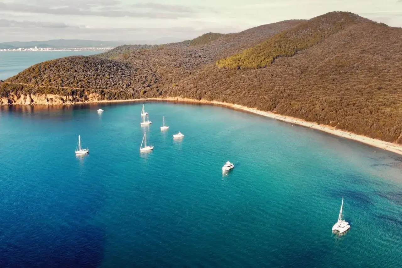 Vue aérienne d'une baie paisible en Maremma, Italie, avec des voiliers et un yacht naviguant sur des eaux turquoise. La côte est bordée d'une plage de sable et de collines verdoyantes.