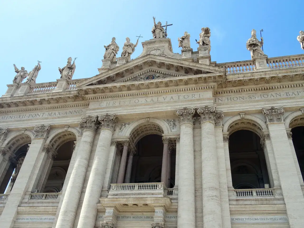 Façade imposante de Saint-Jean-de-Latran, ornée de statues et de colonnes majestueuses sous un ciel bleu.