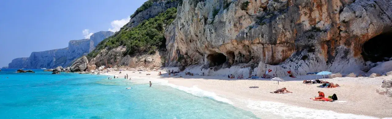 Plage de galets blancs en Sardaigne, avec des falaises imposantes et une eau turquoise. Des baigneurs profitent du soleil, certains à l'ombre de parasols, d'autres se prélassent sur le sable. La ciaccia sardaigne, un paradis naturel.