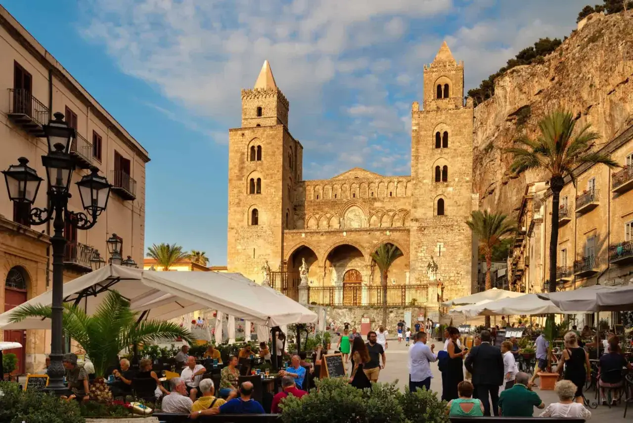 Terrasse animée devant la cathédrale normande de Cefalù, en Sicile. Des palmiers et des bâtiments anciens bordent la place.