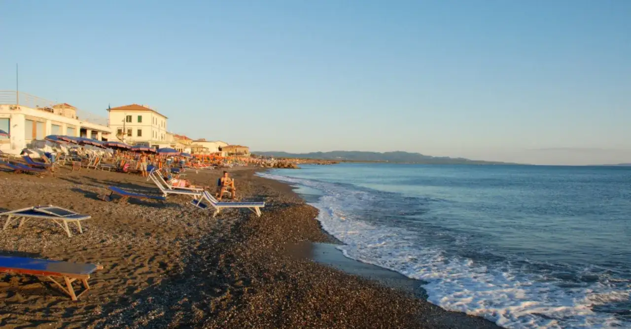 Plage de Cecina Mare au coucher du soleil, avec des chaises longues, des parasols et des bâtiments en bord de mer. L'eau bleue calme rencontre le rivage.