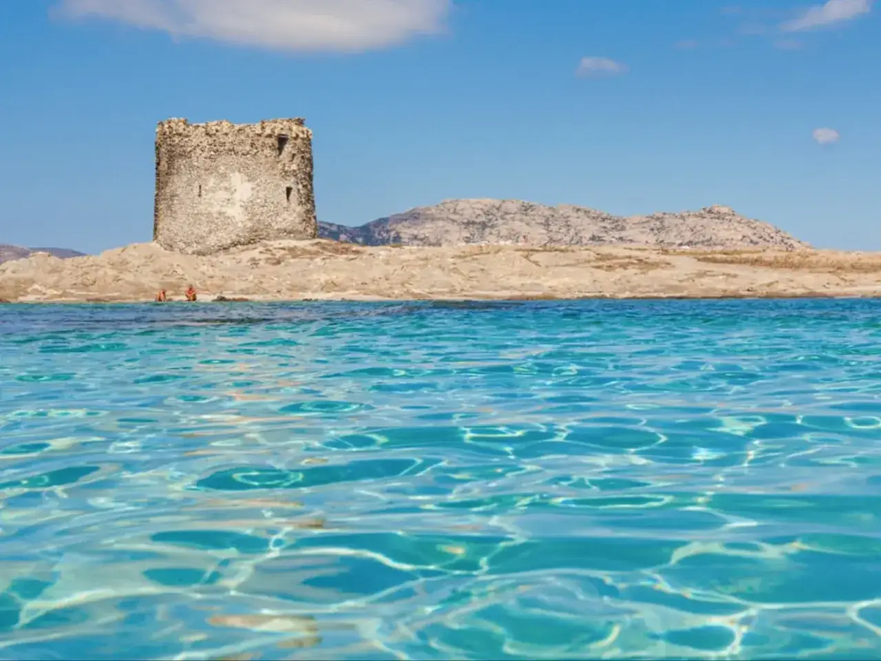 Vue sur une tour de guet en pierre sur une île rocheuse, avec des eaux turquoise claires et des montagnes en arrière-plan. Un aperçu de la plage sud de l'Italie.
