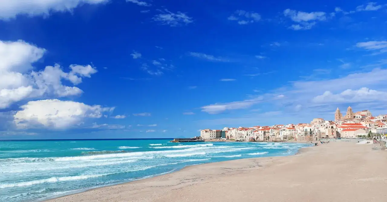 Plage de sable fin, mer turquoise et maisons blanches de Cefalù en Sicile sous un ciel bleu éclatant.
