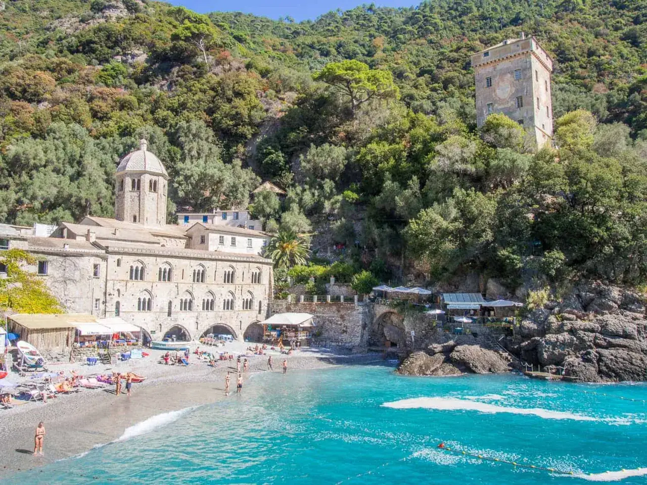 Plage de galets à Rapallo, avec une église médiévale et une tour sur fond de colline verdoyante.