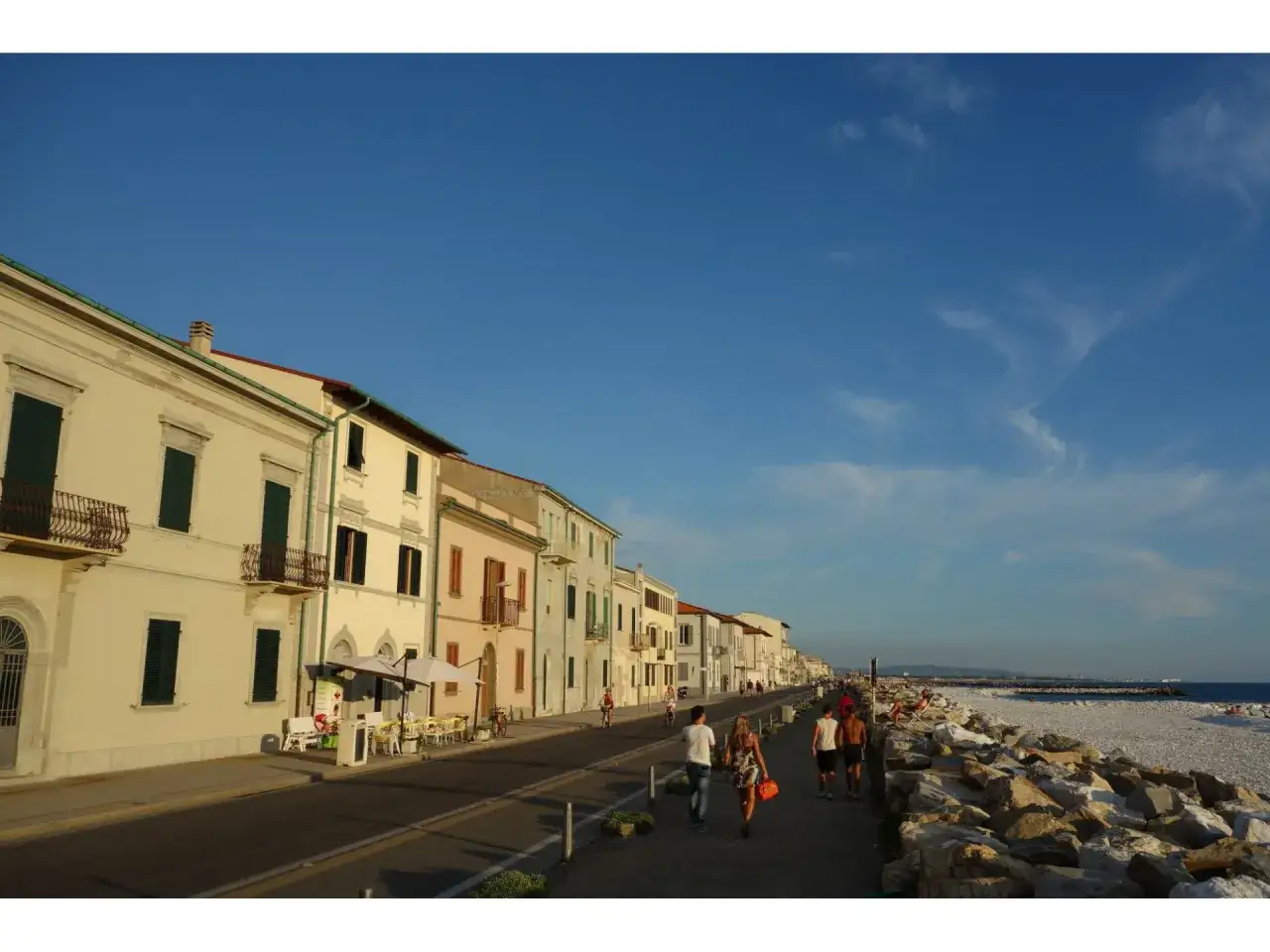Promenade animée le long de la marina de Pise, avec des bâtiments colorés, des cafés en terrasse et des gens se promenant au bord de la mer.