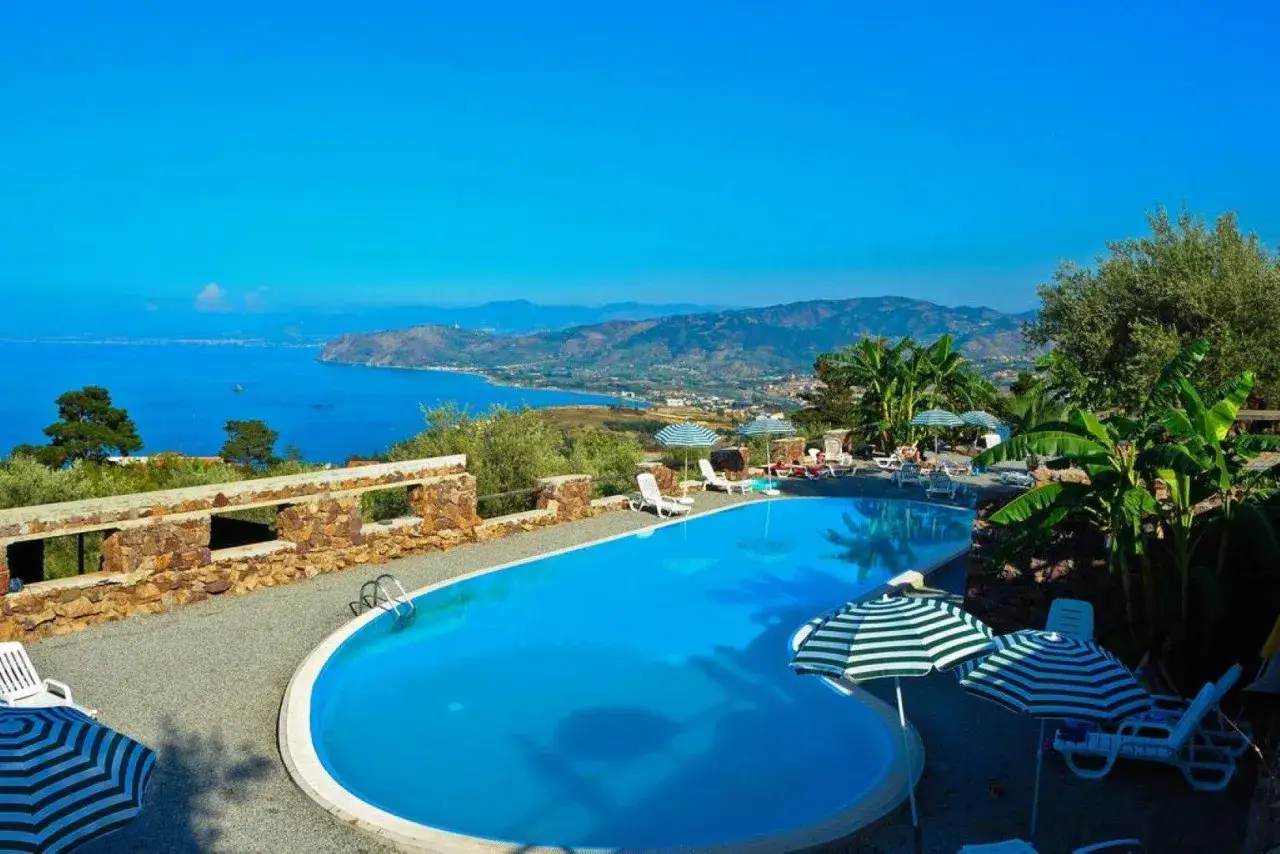 Piscine d'un agroturismo en Sicile, avec vue sur la mer et les montagnes. Chaises longues et parasols invitent à la détente.