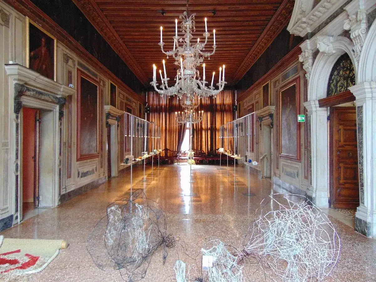 Intérieur du Palazzo Mocenigo, un long couloir orné de tableaux, avec un lustre imposant et des sculptures modernes au sol.