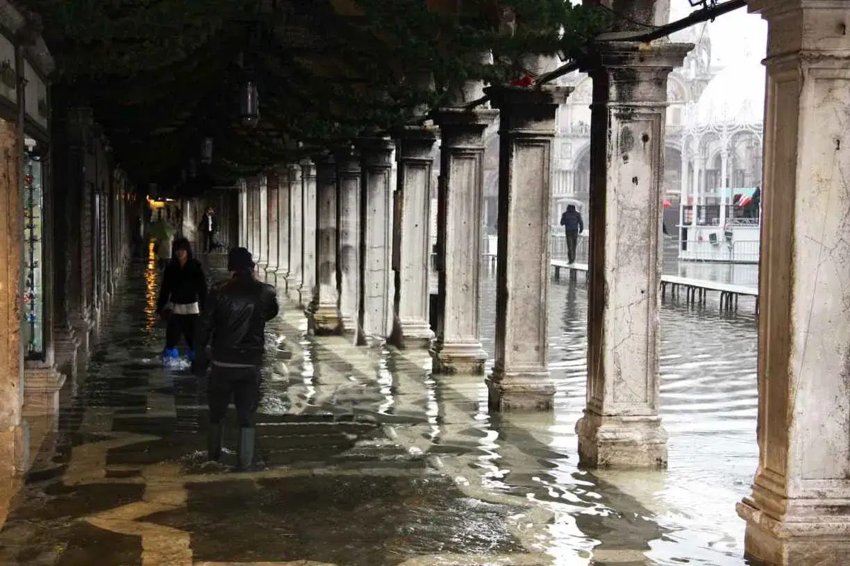 Marée haute à Venise en décembre. Des passants naviguent dans les eaux sous les arcades, reflétant les colonnes imposantes.