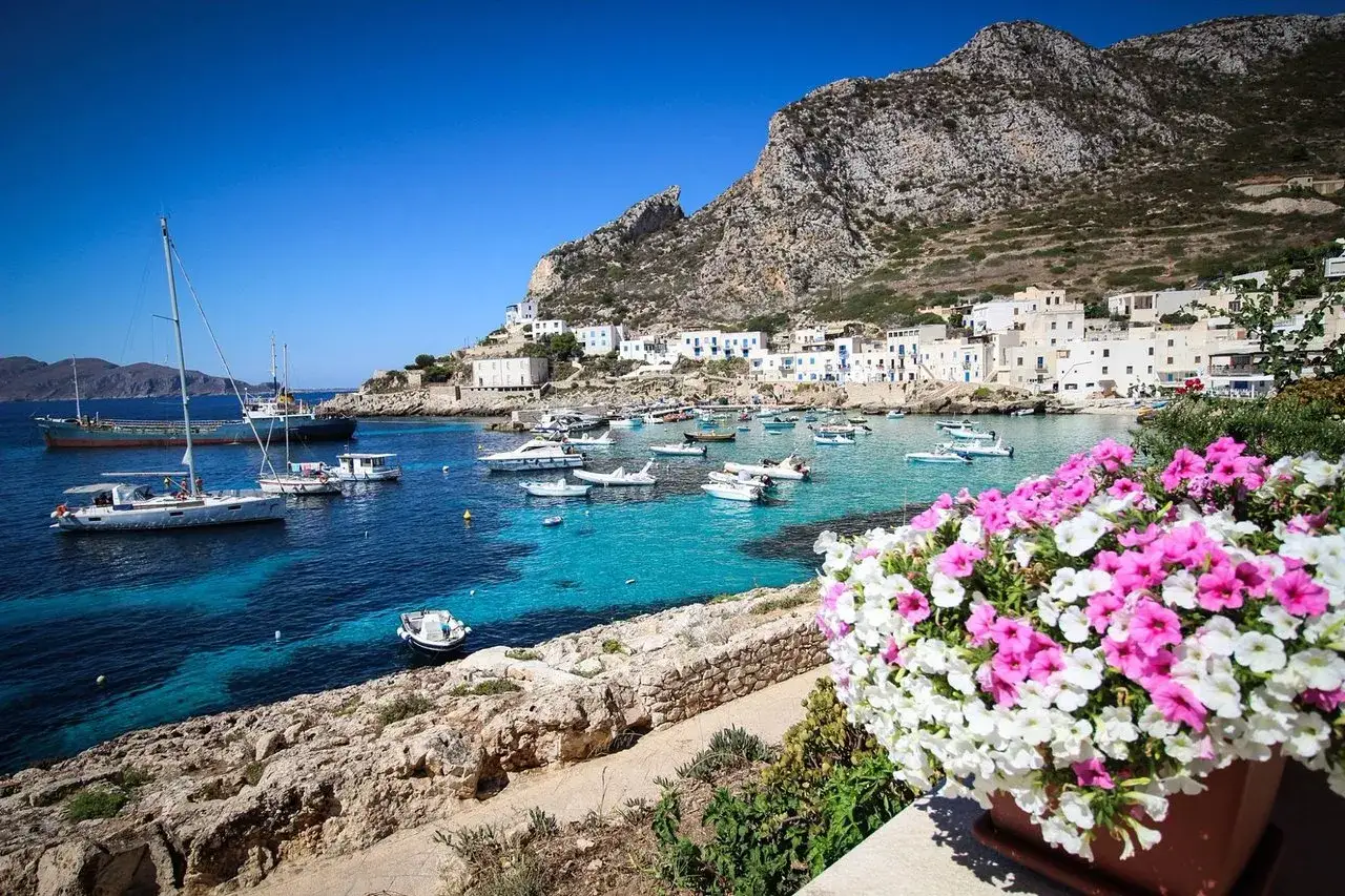 Vue d'un port pittoresque avec des bateaux dans des eaux turquoise, des maisons blanches accrochées à flanc de colline et des fleurs roses et blanches au premier plan. Un coin de paradis en Marsala, Italie.