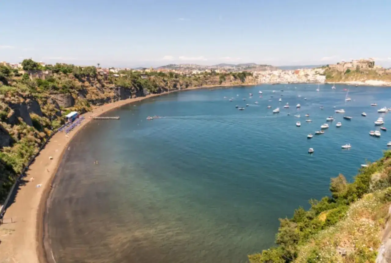 Vue panoramique d'une baie avec une plage de sable noir, des falaises verdoyantes et de nombreux bateaux dans l'eau bleue. On aperçoit une ville colorée au loin, évoquant l'atmosphère de Procida ou Capri.