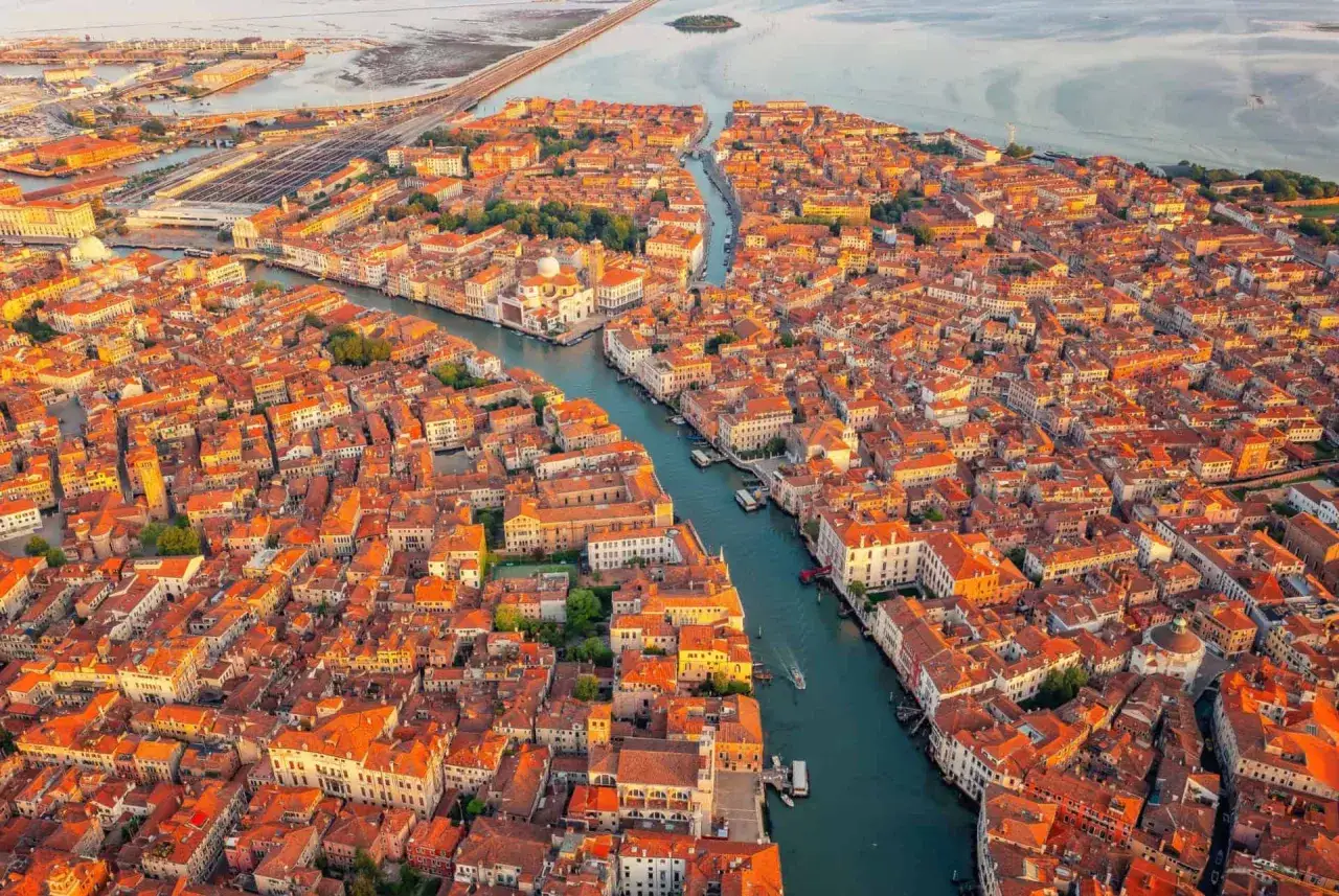 Vue aérienne de Venise au coucher du soleil, avec ses canaux sinueux et ses toits orangés. Un pont emblématique de Venise se dessine au loin.
