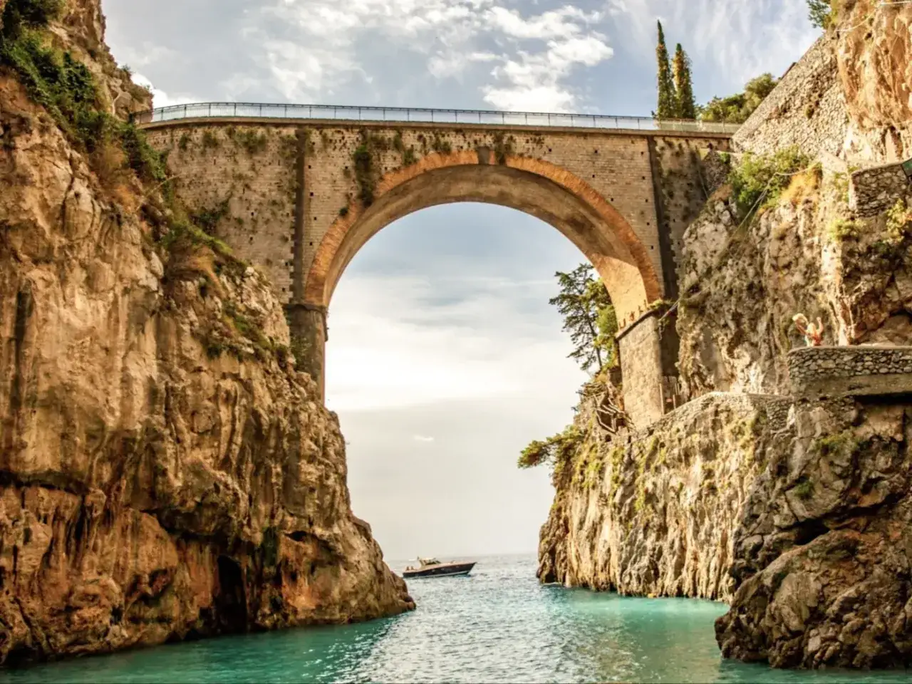 Un pont majestueux enjambe une crique aux eaux turquoise, typique de la côte amalfitaine en Italie. Un bateau navigue paisiblement.