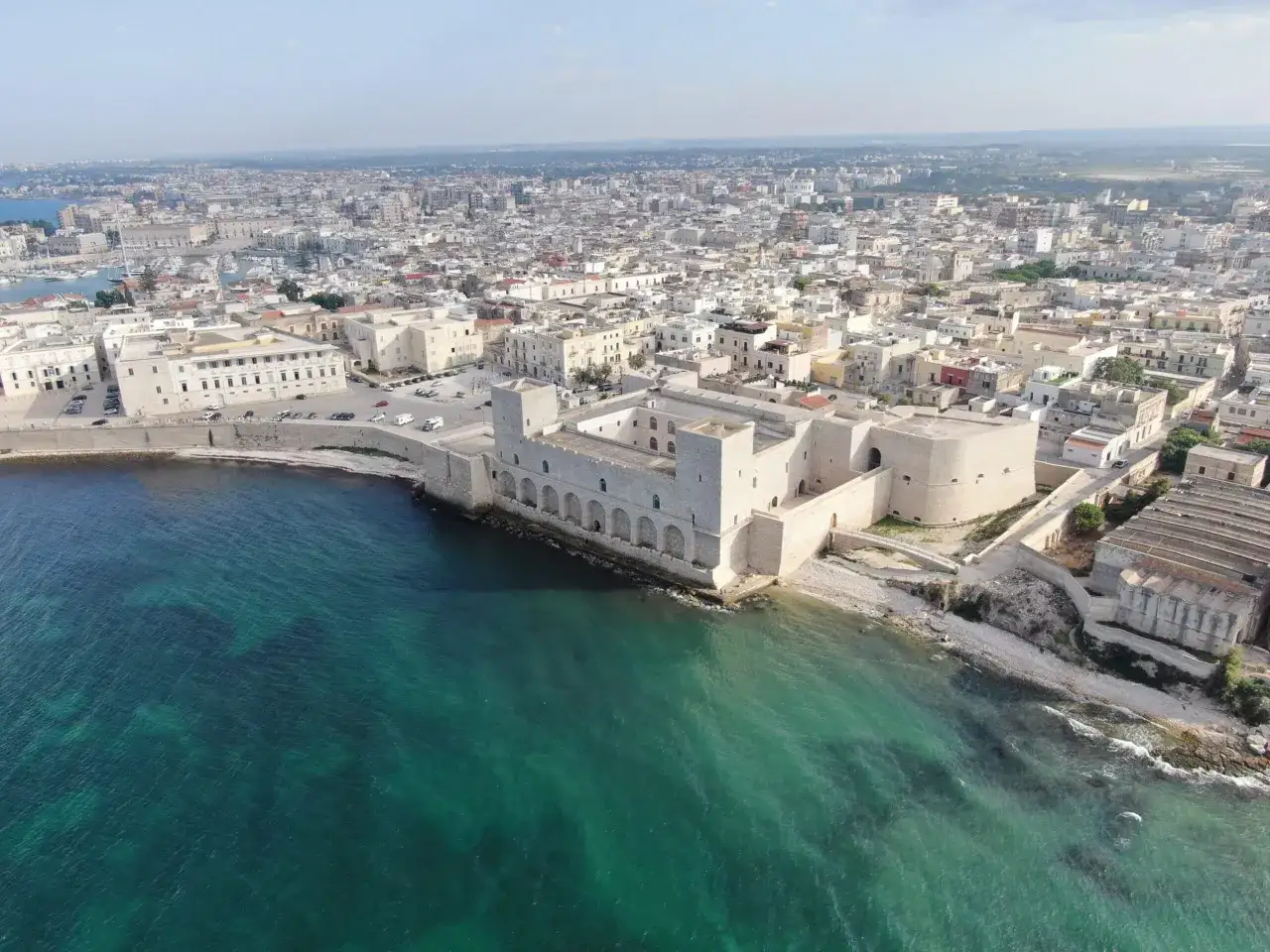 Vue aérienne du château de Trani, une forteresse médiévale sur la côte, avec la ville en arrière-plan et la mer d'un bleu-vert éclatant.