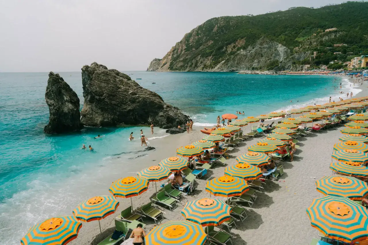Plage animée de la riviera italienne où aller pour se baigner et se détendre sous des parasols colorés, avec des rochers imposants et une mer turquoise.
