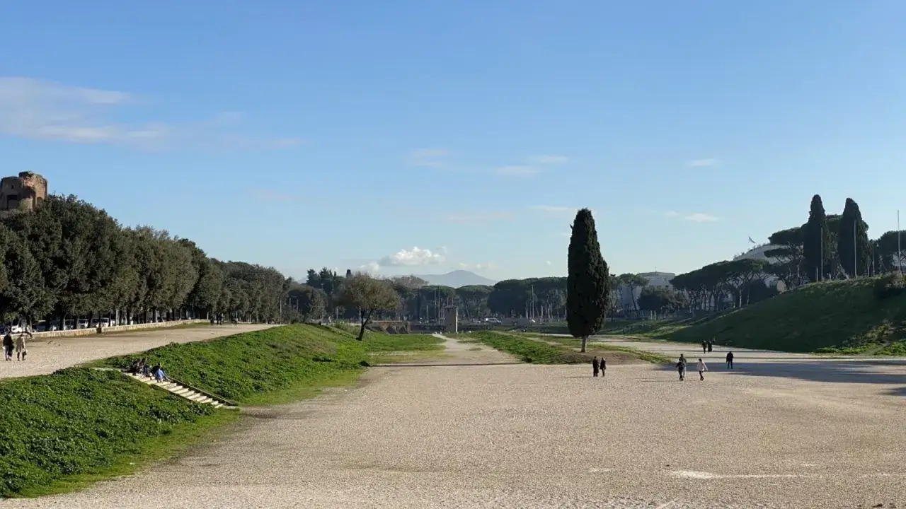 Vaste étendue de gravier au Circo Massimo à Rome, bordée d'arbres et de verdure. Quelques personnes se promènent sous un ciel bleu clair.