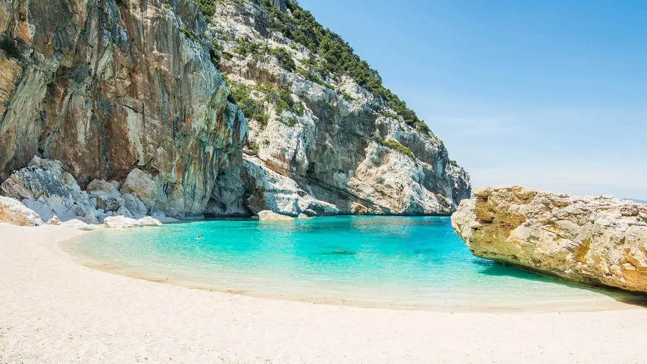 Plage de sable blanc bordée d'eaux turquoise cristallines, avec d'imposantes falaises rocheuses verdoyantes en arrière-plan. Un paradis baigné de soleil, digne de Portonovo.