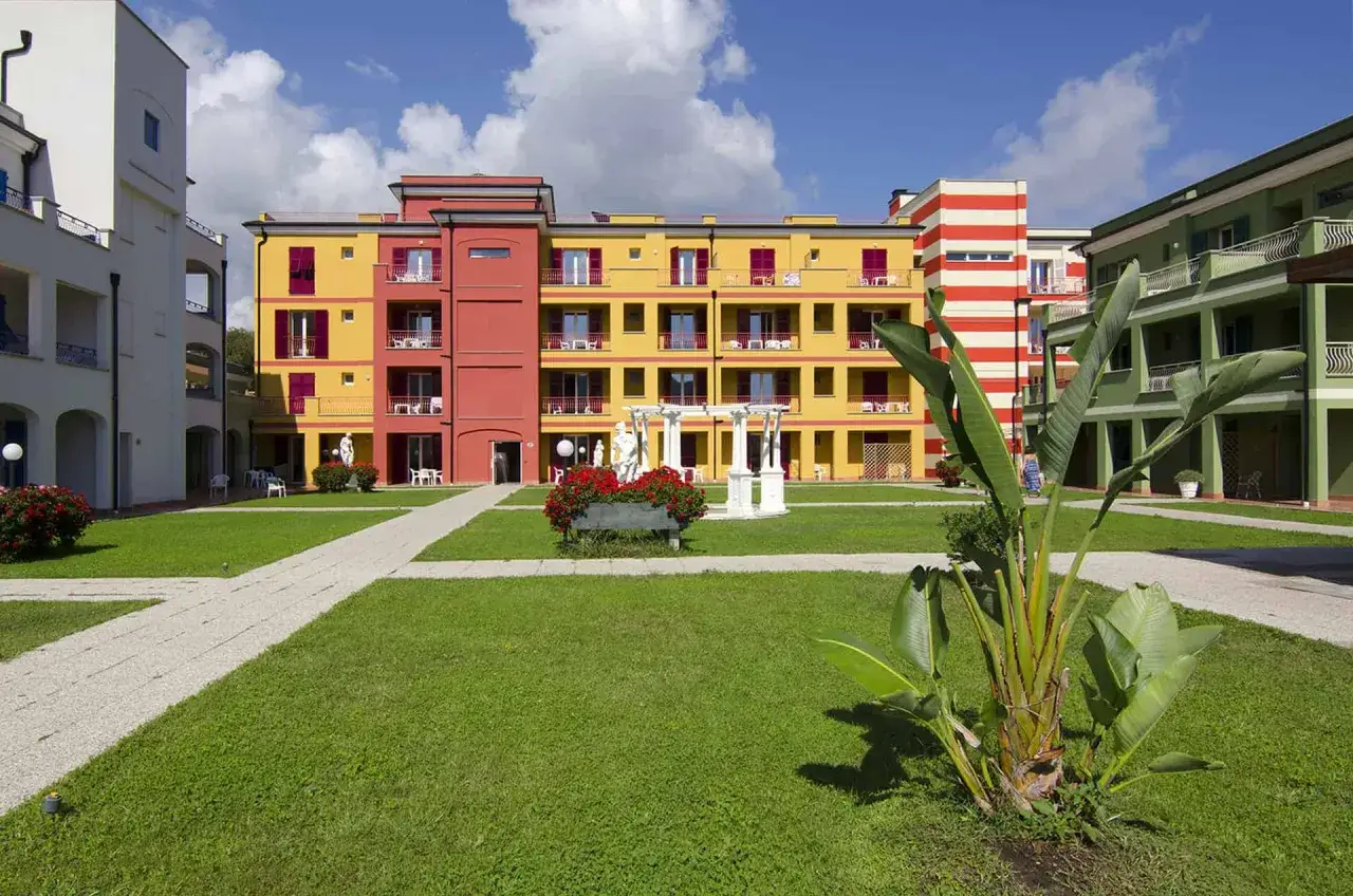 Cour int&eacute;rieure color&eacute;e du village Ai Pozzi, avec des b&acirc;timents jaune et rouge, une pelouse verte et des plantes tropicales.