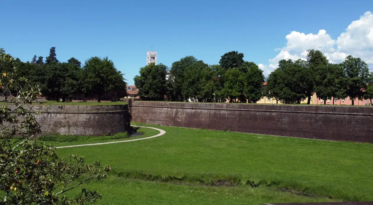 Murs de Lucques, avec une tour m&eacute;di&eacute;vale visible derri&egrave;re les arbres. Un chemin serpente dans l'herbe verte.