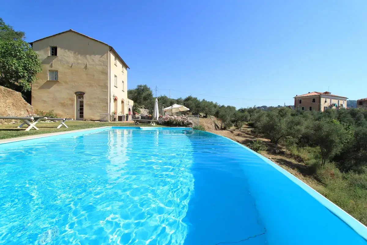 Piscine étincelante d'un village vacances à La Spezia, avec une maison toscane et des oliviers en arrière-plan.