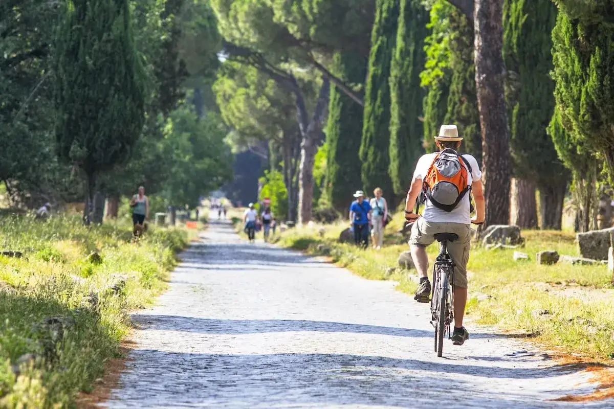 Un homme à vélo explore la voie Appienne, un site historique près de Rome, entouré de cyprès et de ruines antiques.